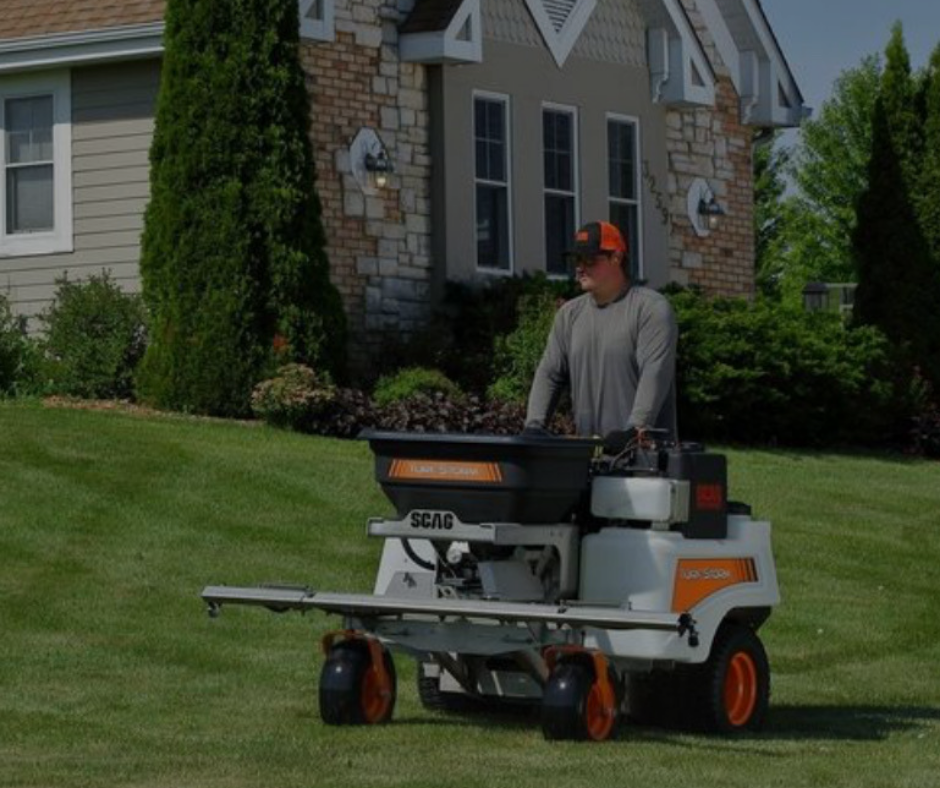Man operating a lawn spreader on a residential lawn. The spreader is white and orange.