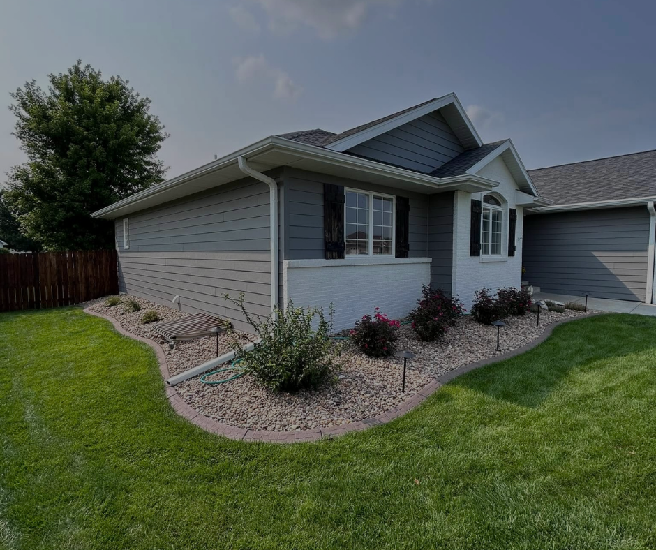 A ranch-style house with gray siding, a white trim, and a well-manicured lawn and landscaping under a blue sky.