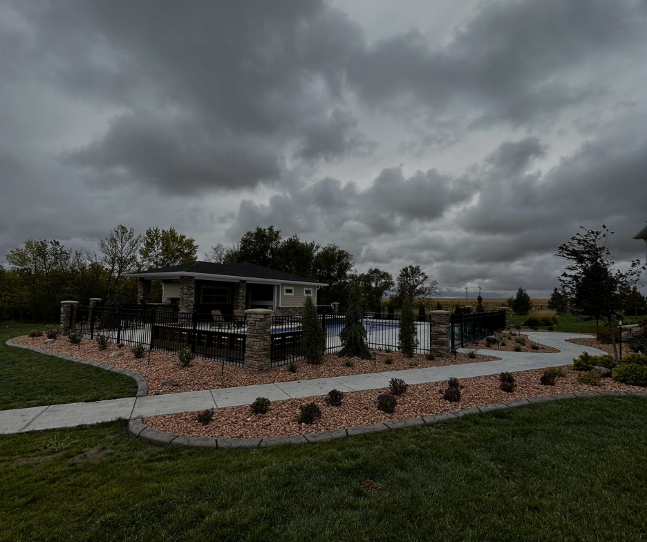 Poolside cabana under a cloudy sky, with a paved path, landscaping, and green grass.