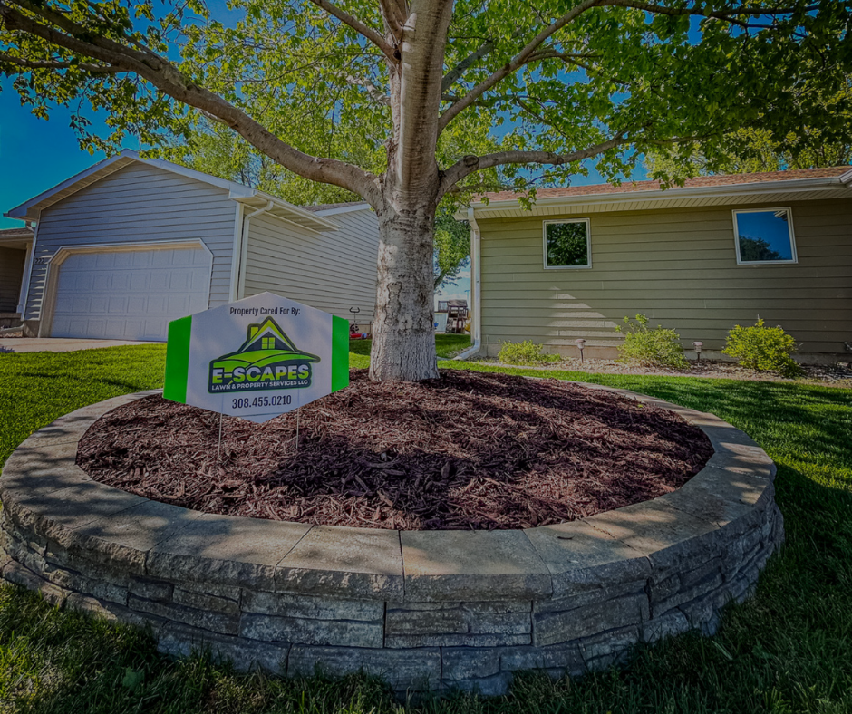 Landscaped tree bed with retaining wall, mulch, and a sign for E-Scaped Landscaping in front of a house.