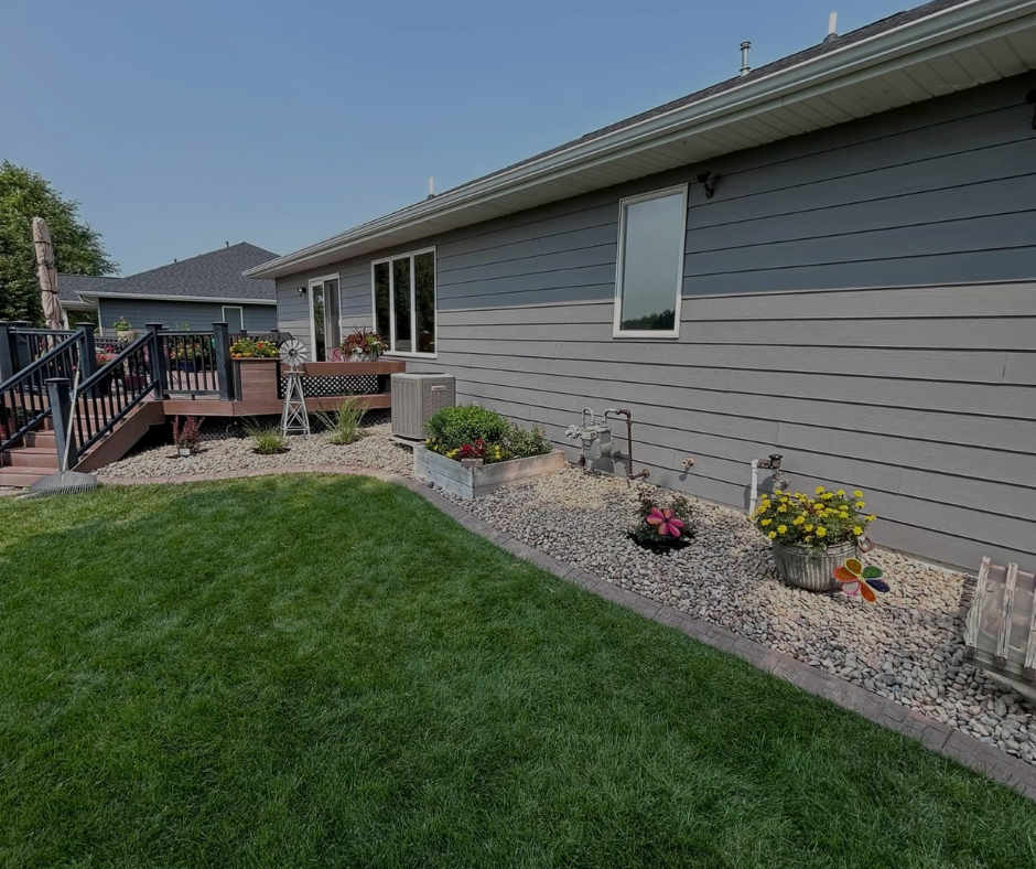 Backyard with gray house, deck, flowerbeds, and green lawn.