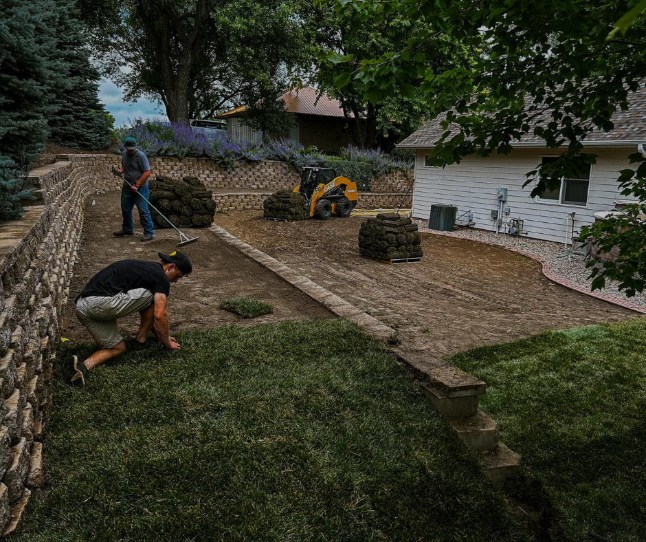 Two men laying sod in a backyard next to a retaining wall and a house. Green grass, cloudy sky.