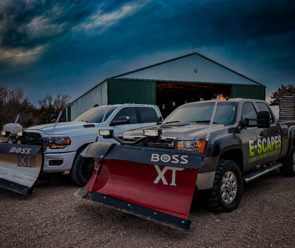 Two snowplow trucks parked in front of a green barn. One white, one dark gray, both with red plows. Cloudy sky.
