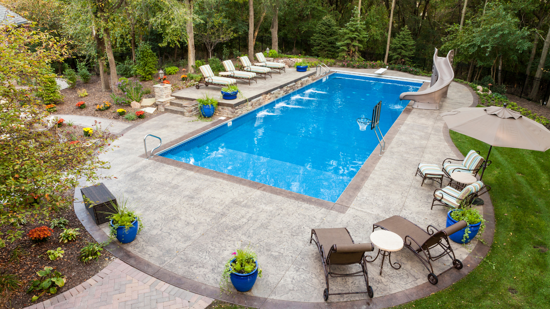 Swimming pool with lounge chairs, slide, and patio area surrounded by greenery.