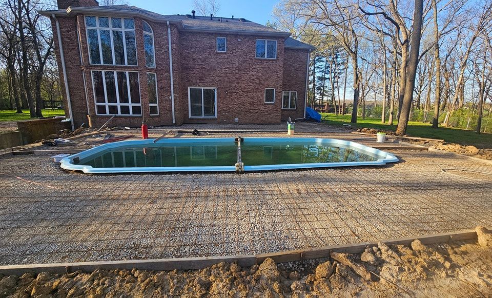 Pool under construction in front of a two-story brick house, gravel, and bare trees surround the area.