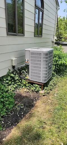 An air conditioning unit next to a light-colored building with windows and overgrown plants.