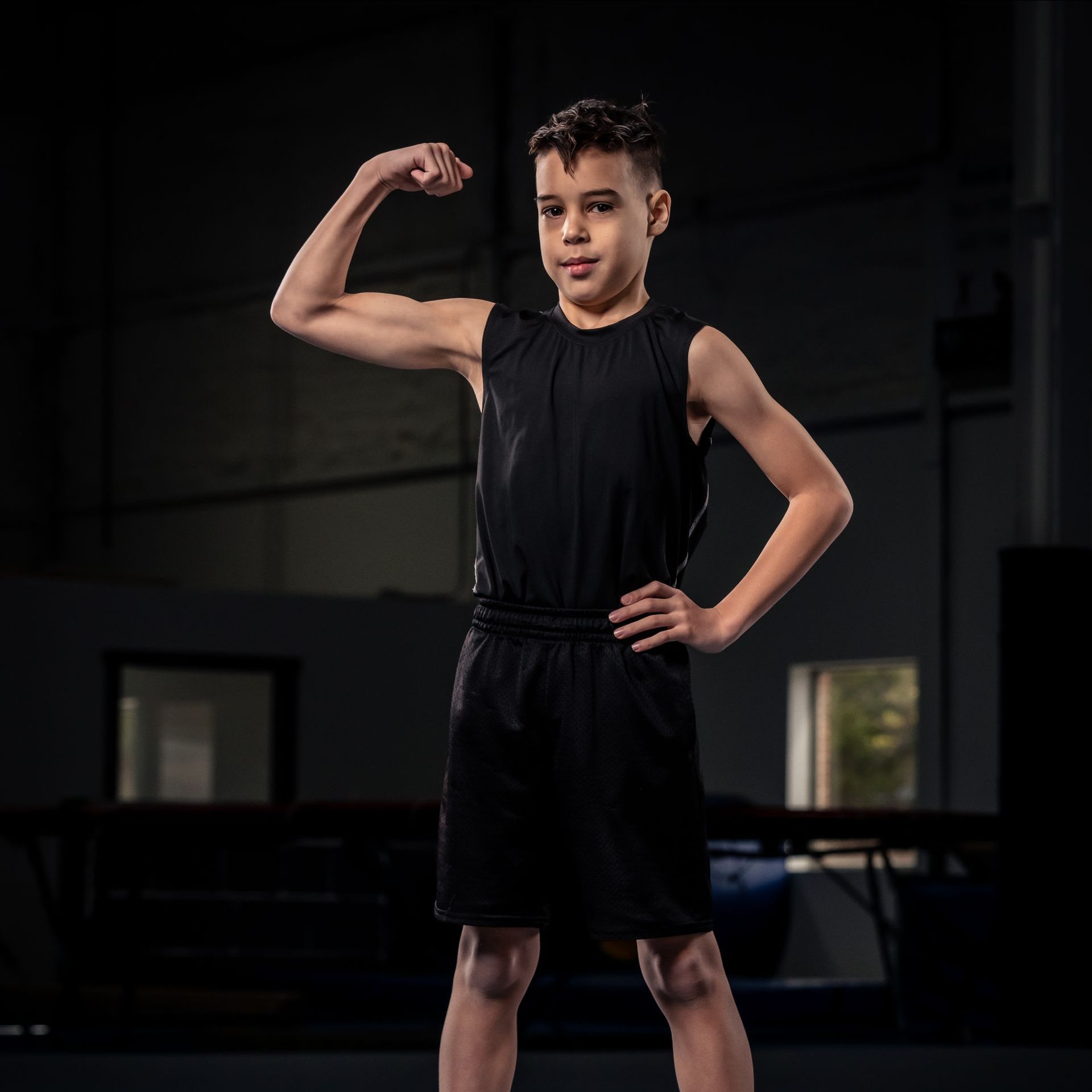 A young boy in a black tank top and shorts is flexing his muscles.