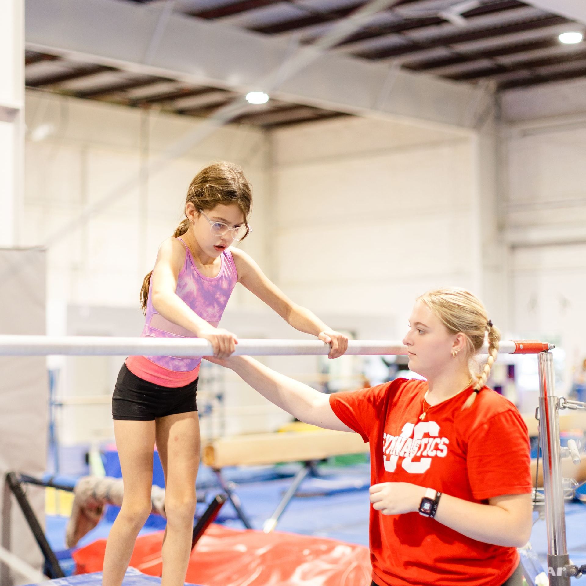 A woman in a red shirt is helping a young girl on a balance beam.