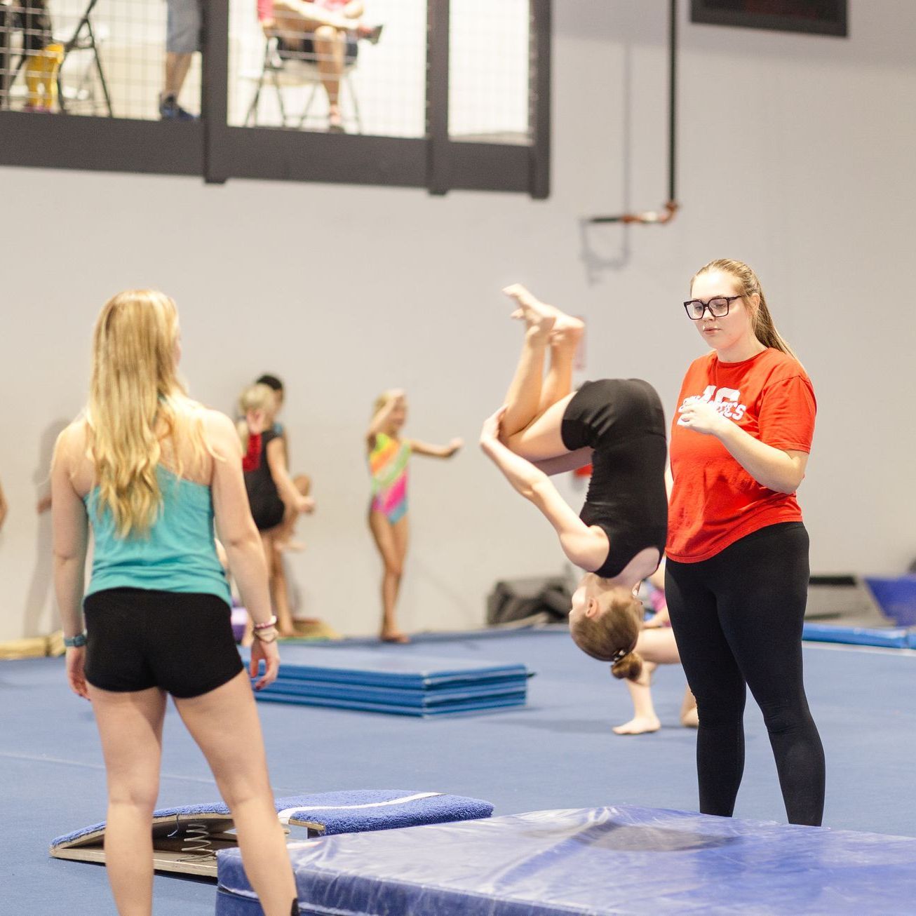 A woman in a red shirt is watching a girl do a handstand on a mat