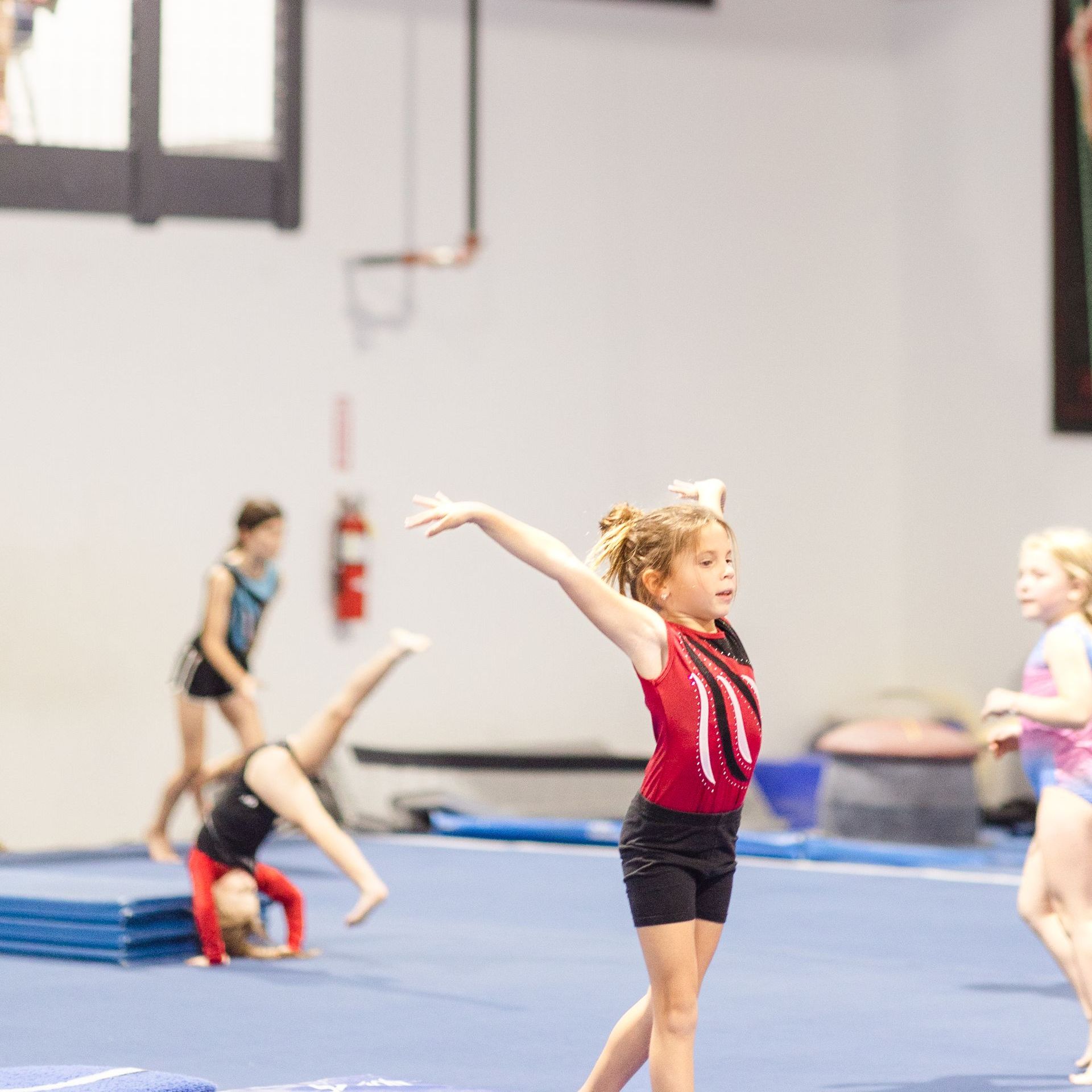 A young girl in a red leotard is doing a handstand on a gym floor.