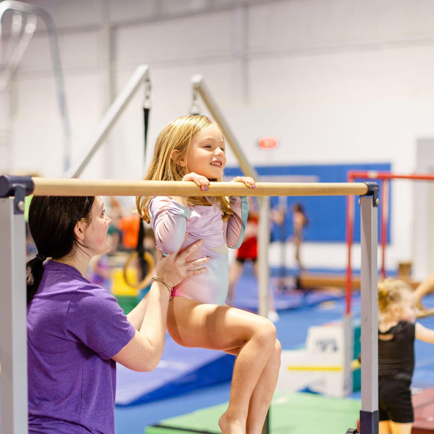 A little girl is hanging from a bar in a gym while a woman watches.