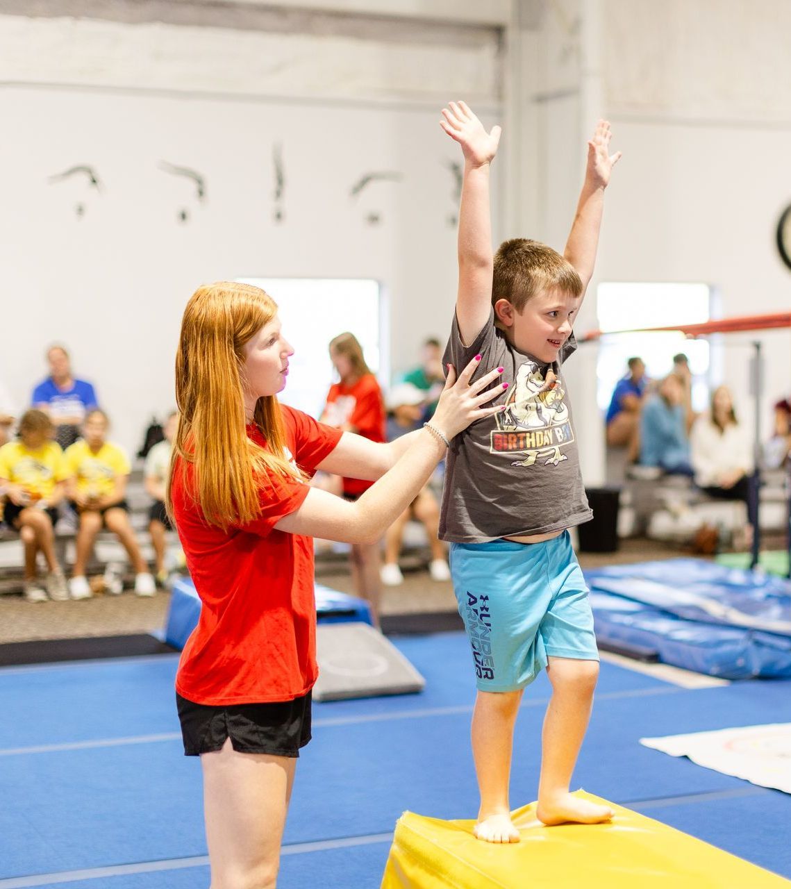 A young boy is standing on a yellow box with his arms in the air while a woman watches.