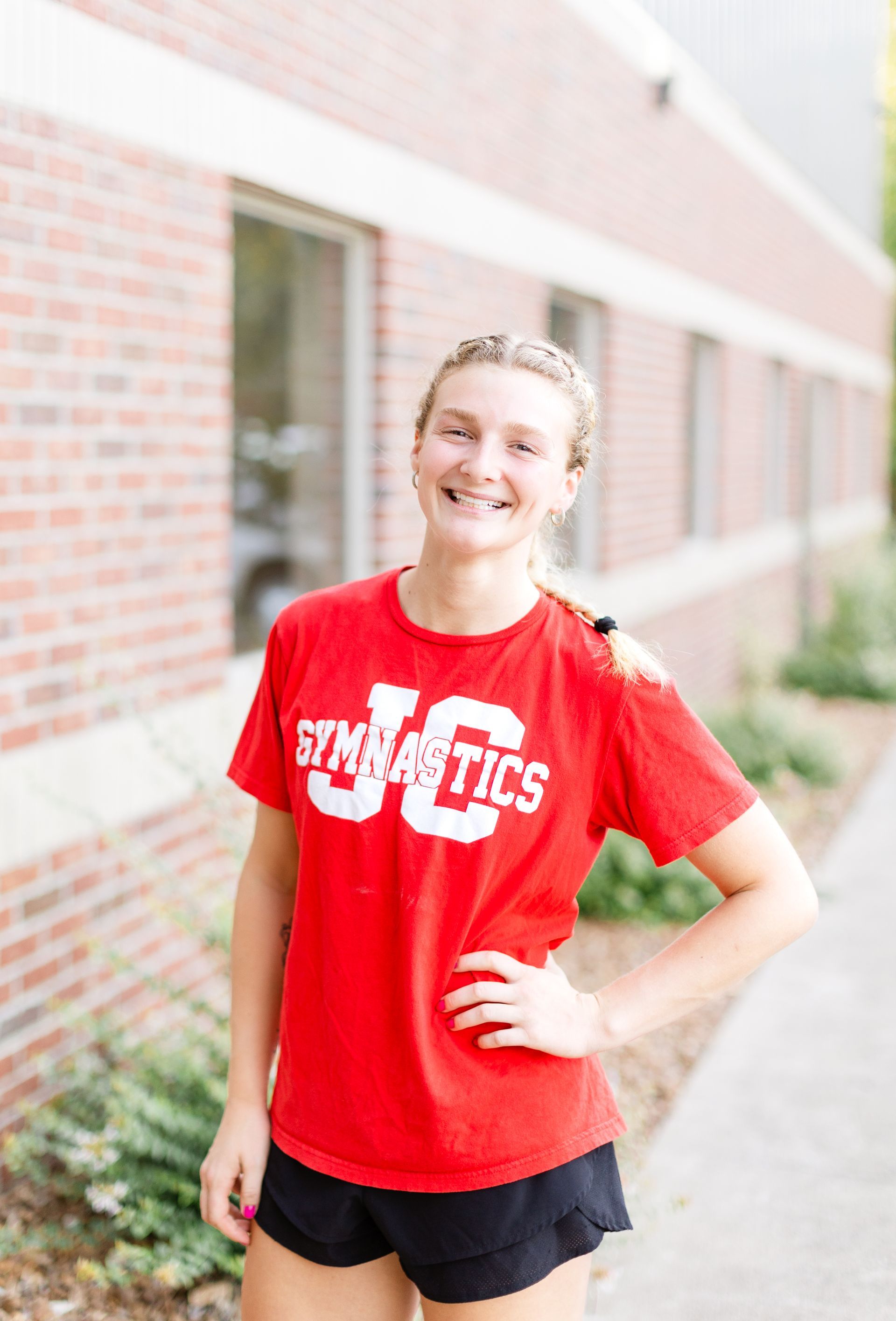 A woman in a red t-shirt is standing in front of a brick building.
