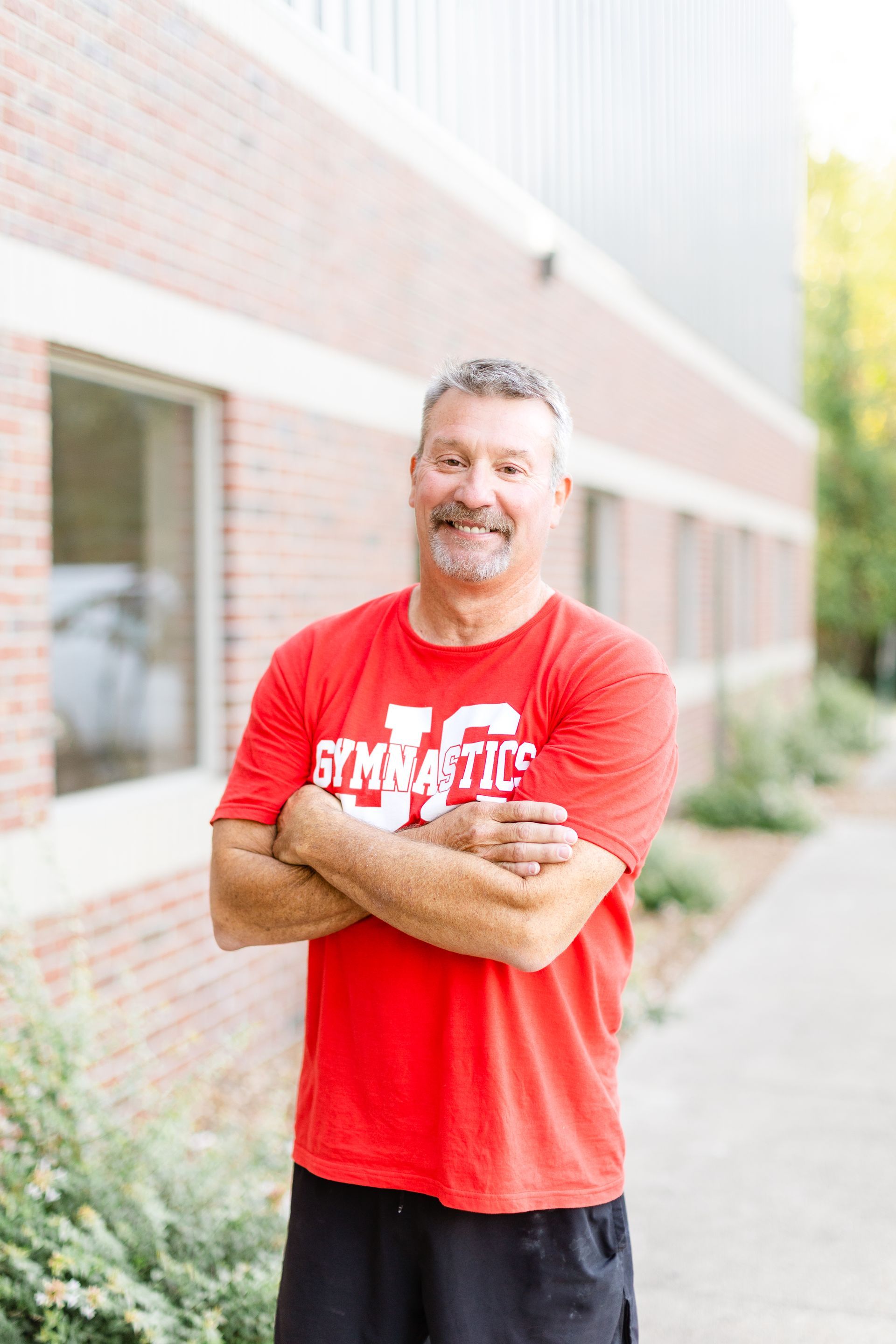 A man in a red shirt is standing with his arms crossed in front of a brick building.