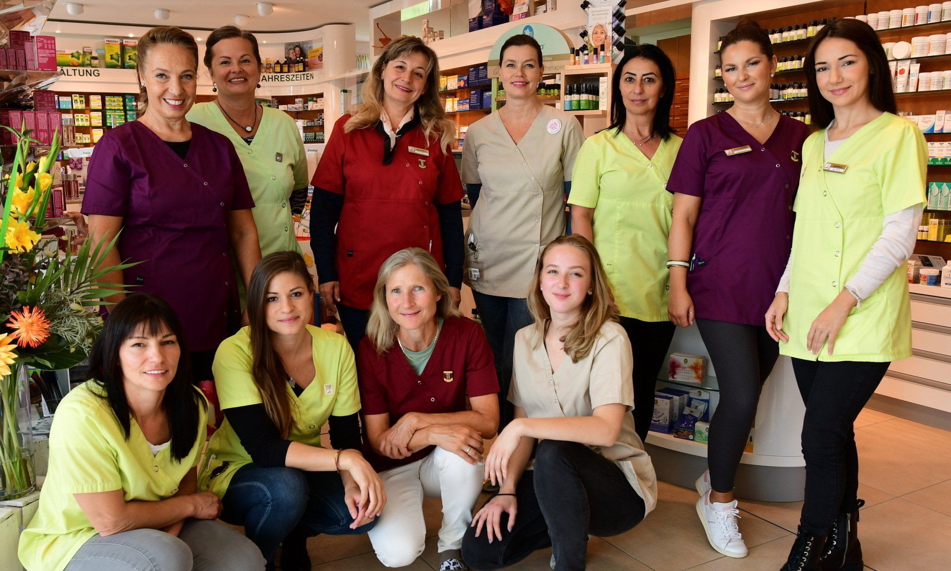 Eine Gruppe von Apothekenmitarbeitern in verschiedenfarbigen OP-Kitteln posieren lächelnd in einem Innenraum.