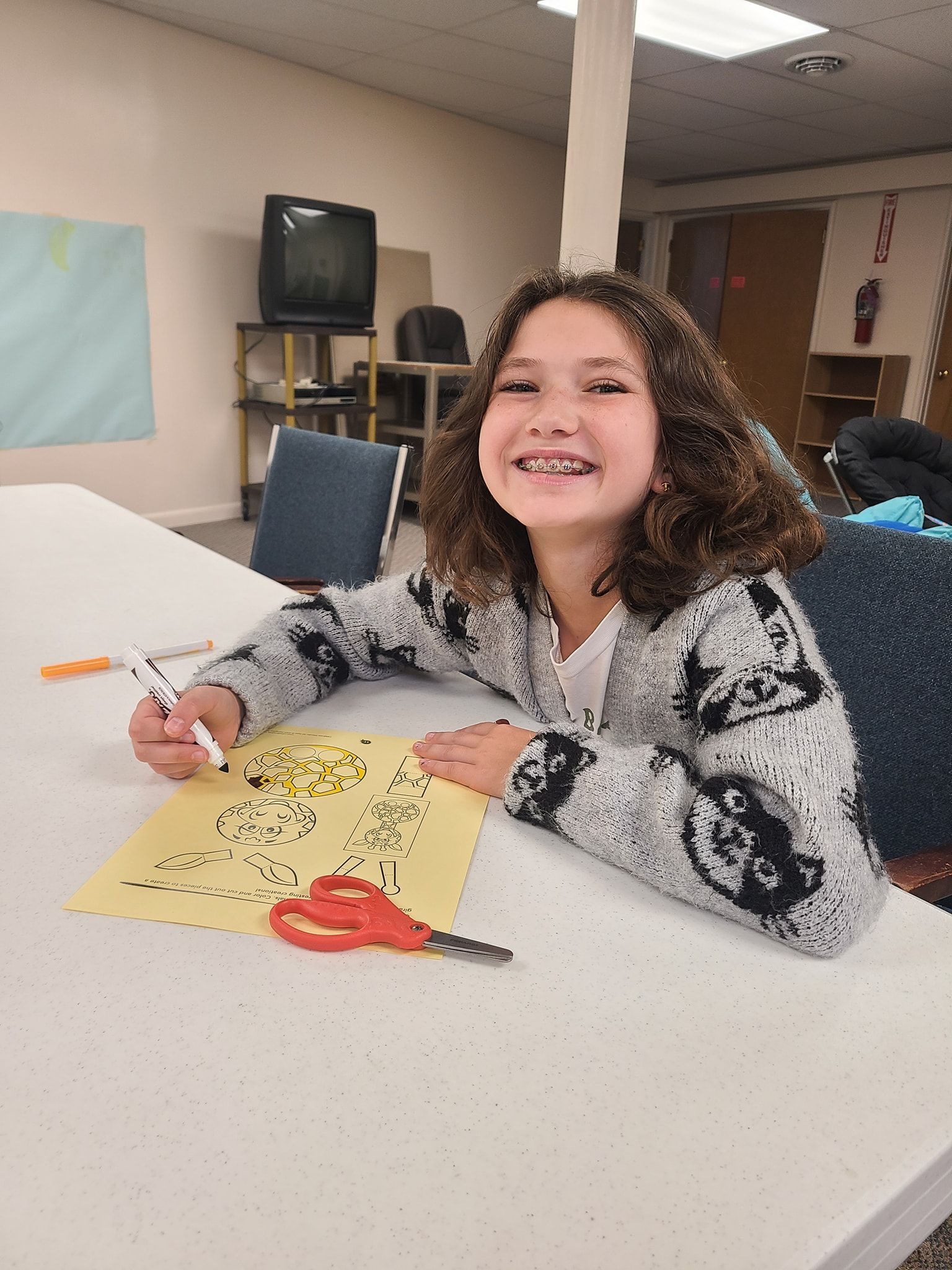 Girl with braces smiles, drawing at a table. She wears a grey sweater with cat faces. Scissors and a yellow paper are visible.