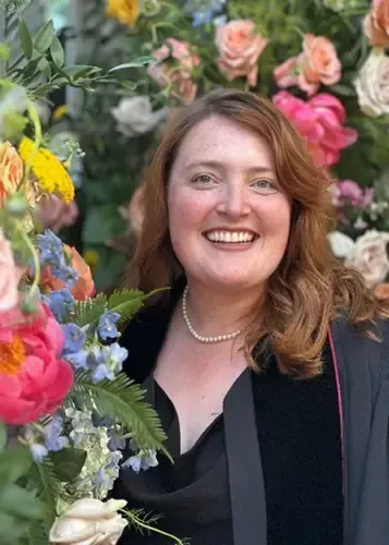 Woman with red hair smiles, wearing a pearl necklace and black graduation attire, in front of colorful flowers.