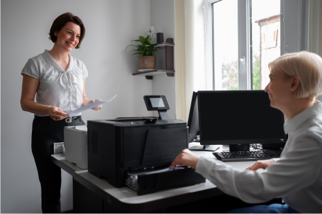 A woman is standing next to a man sitting at a desk in front of a printer.