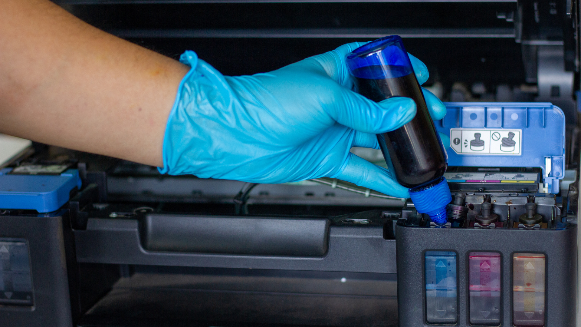 Person refilling blue ink in a printer with a bottle, wearing a blue glove.