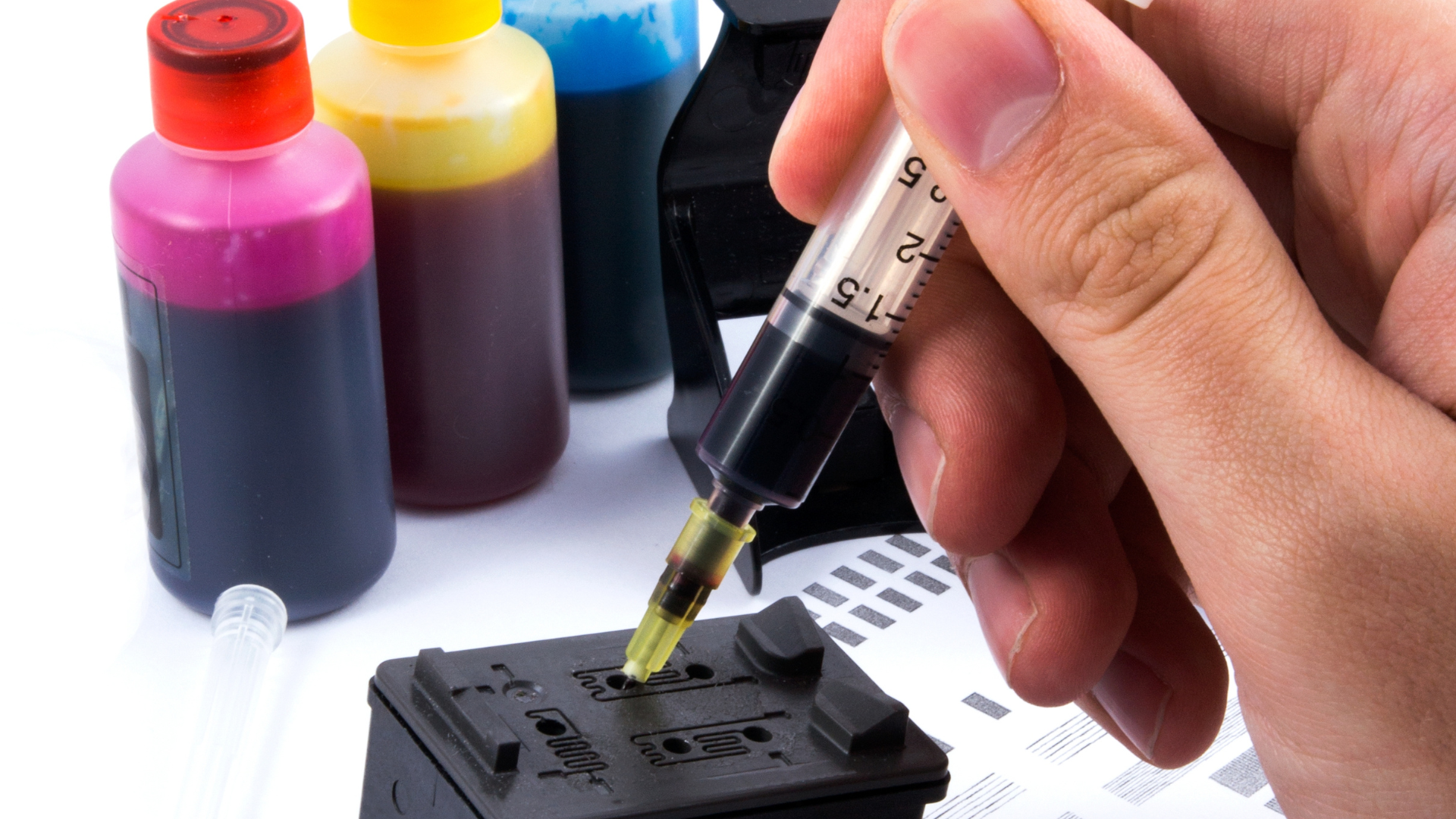 Hand injecting black ink into a printer cartridge with ink bottles in the background.