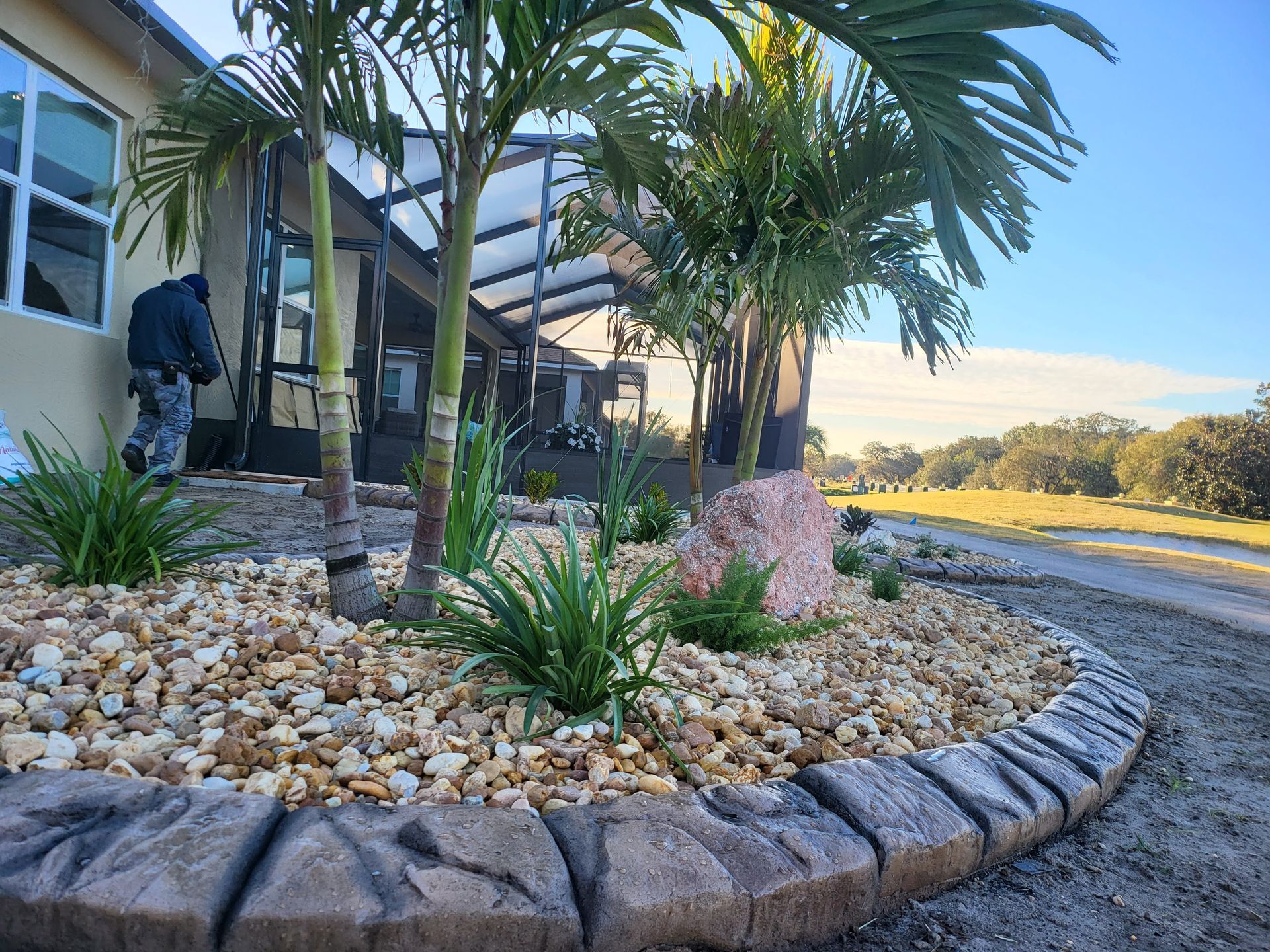 Landscaped flowerbed with palm trees, rocks, and greenery next to a house with a man in a coat.