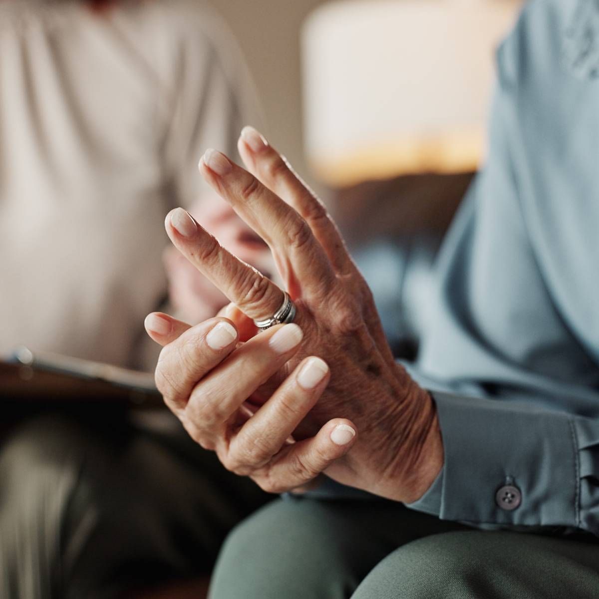 Person's hands, one wearing a ring, touching the other. Blurred person in the background, likely indoors.