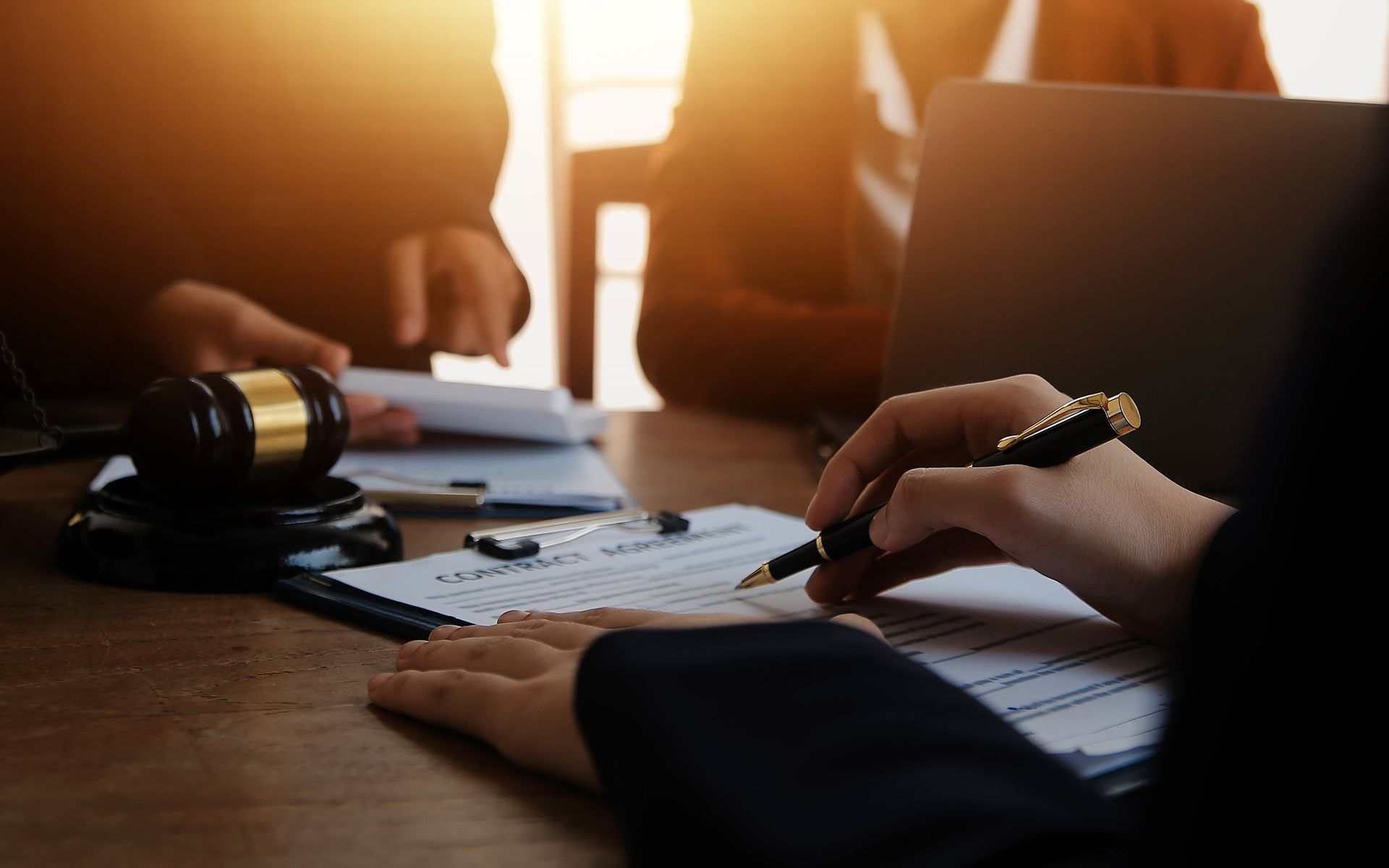 Hands signing a contract at a table with a gavel, papers, and laptop in a room.