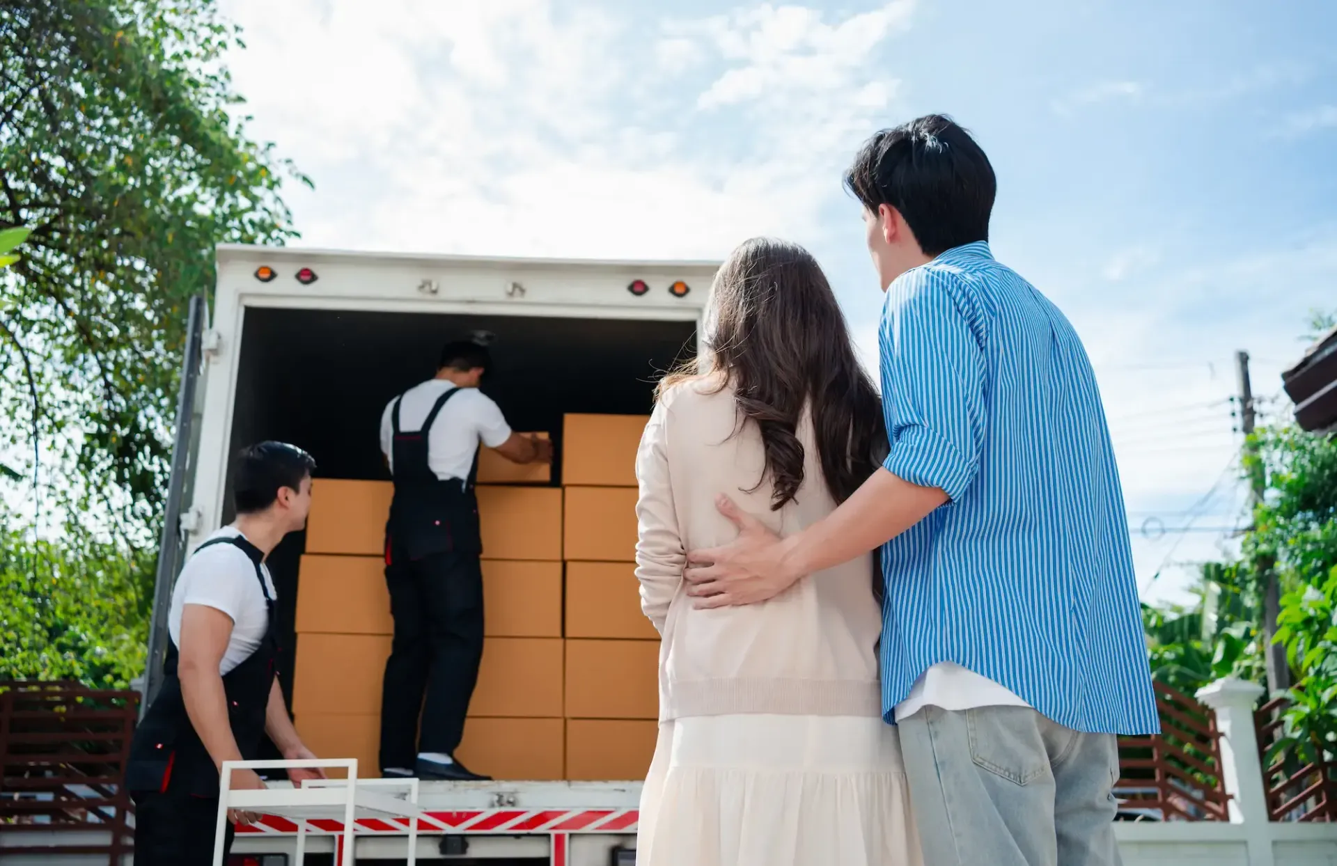 Couple watching movers load boxes into a truck on a sunny day.