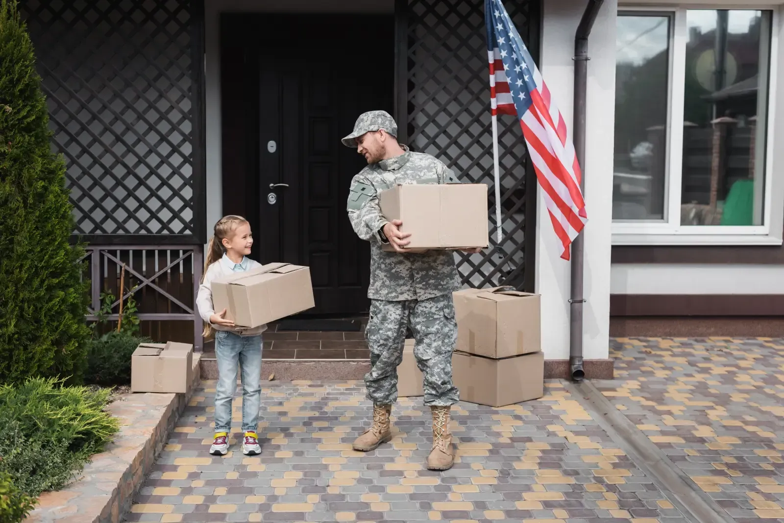 A soldier in uniform and a young girl carry cardboard boxes near a house with an American flag.