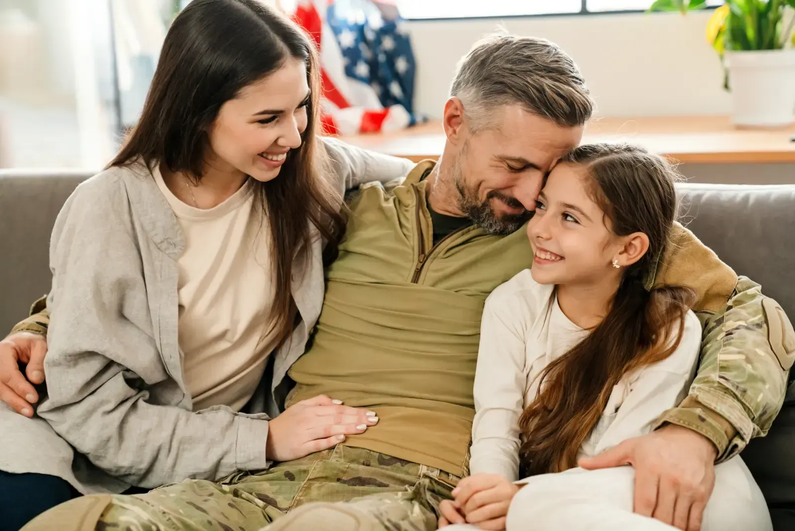 A military service member in uniform sits on a couch, embracing a person and a child who are smiling at them.