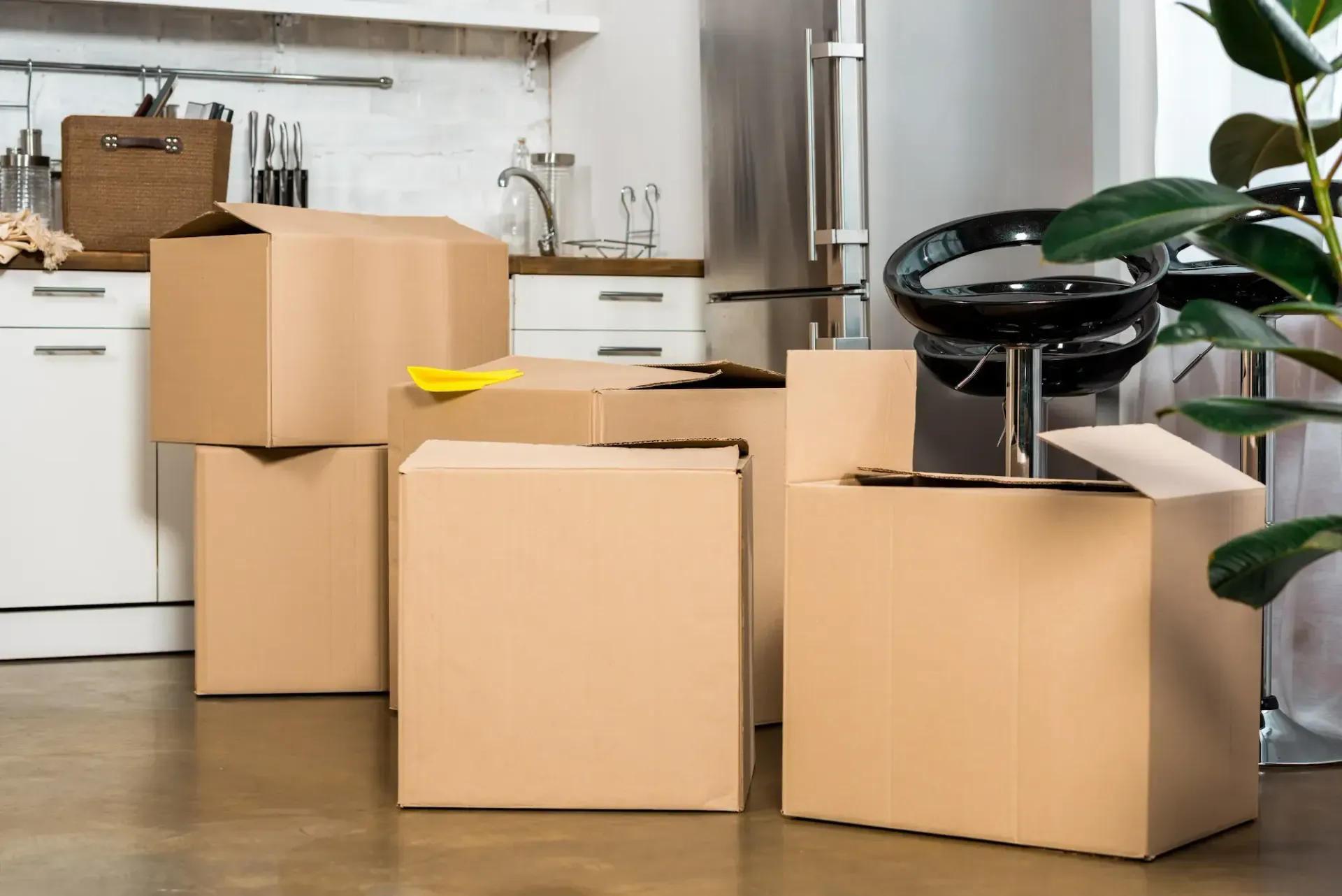 Cardboard moving boxes stacked in a kitchen near a stainless steel refrigerator and a bar stool.