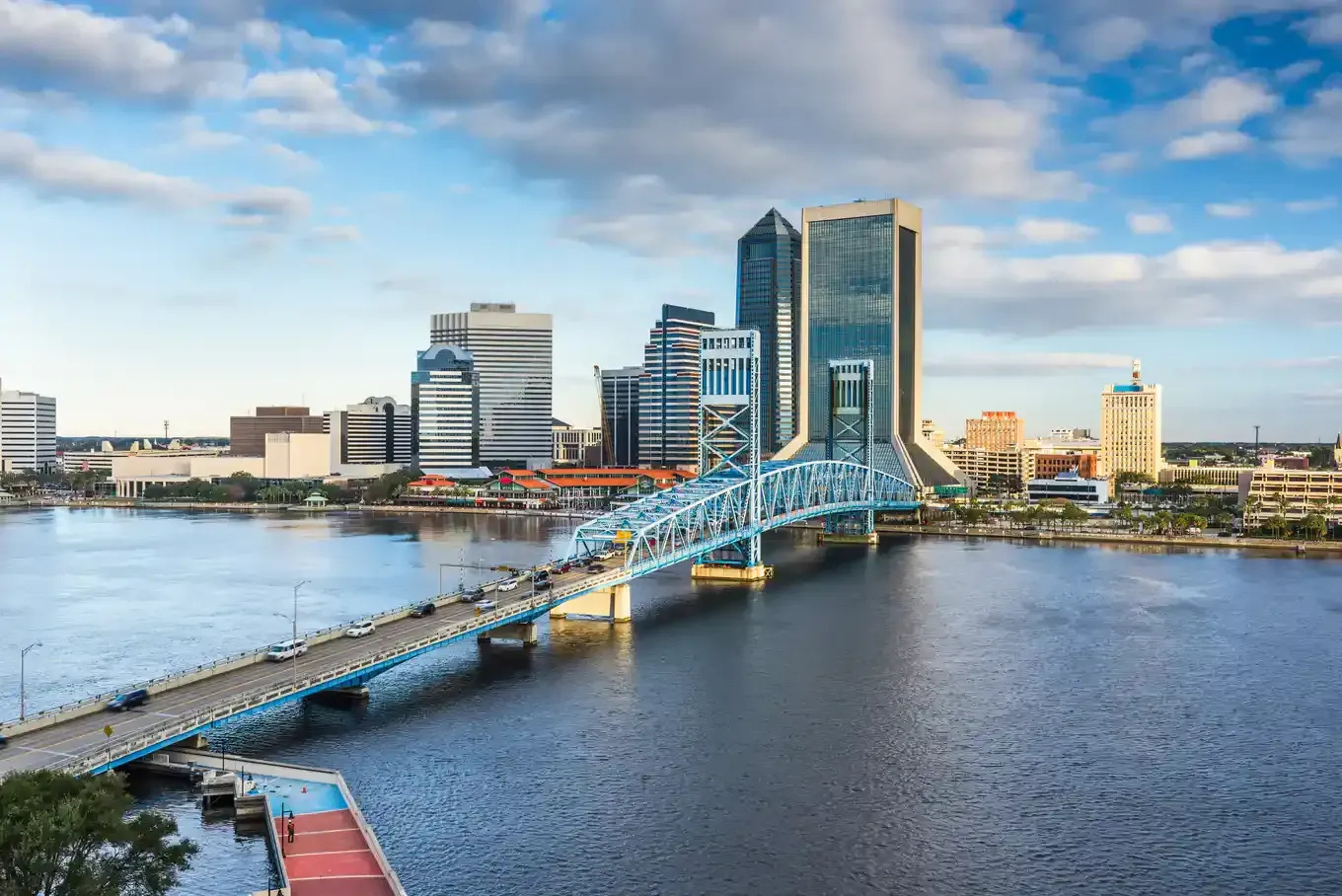 Blue bridge spanning a river, Jacksonville, Florida skyline in background, under cloudy sky.