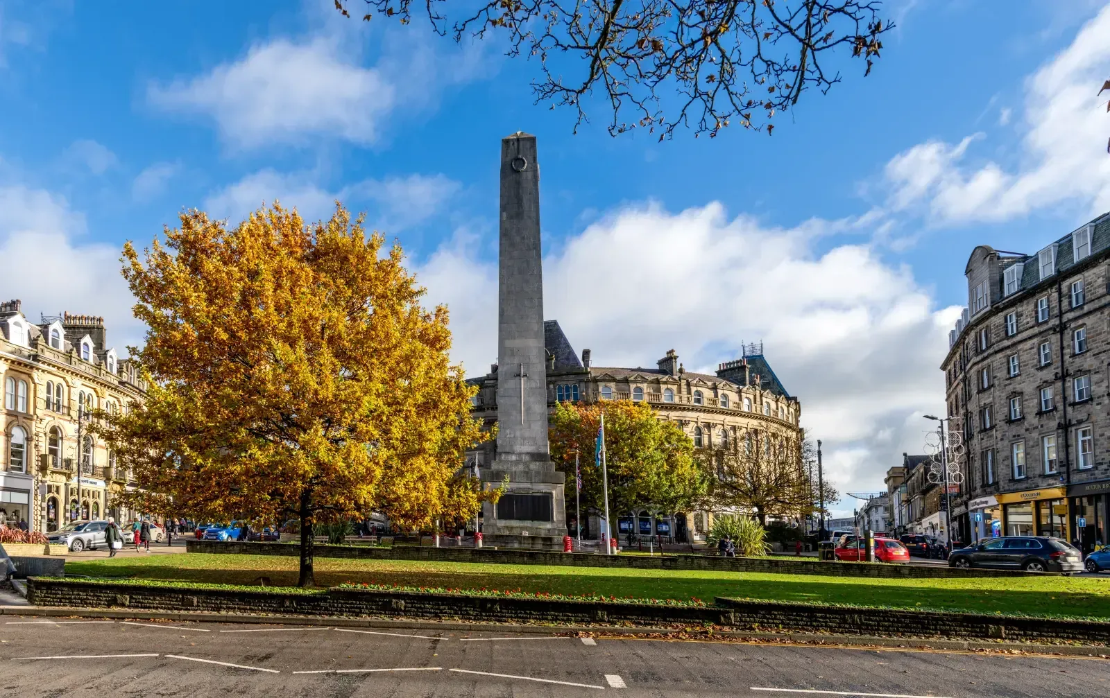 A tall stone war memorial stands in a green town square park with a large golden-leaved tree under a bright blue sky.