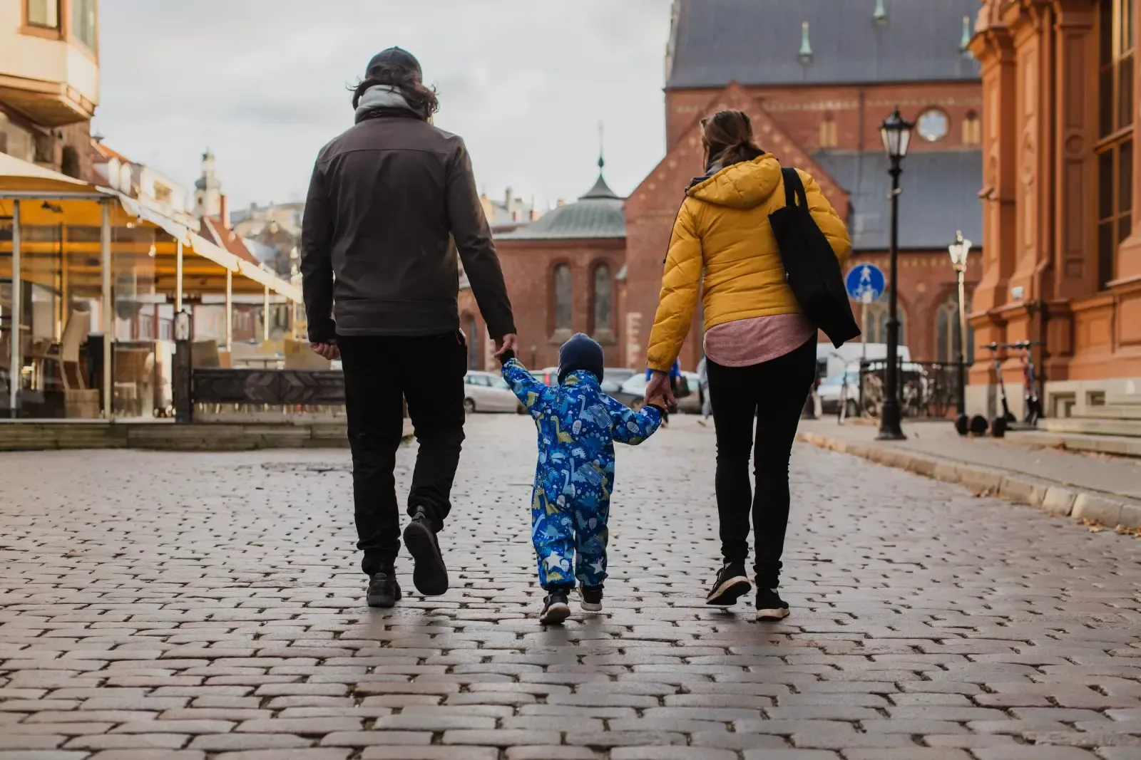 A parent in a dark jacket and one in a yellow puffer jacket hold hands with a toddler in a blue snowsuit on a cobblestone.