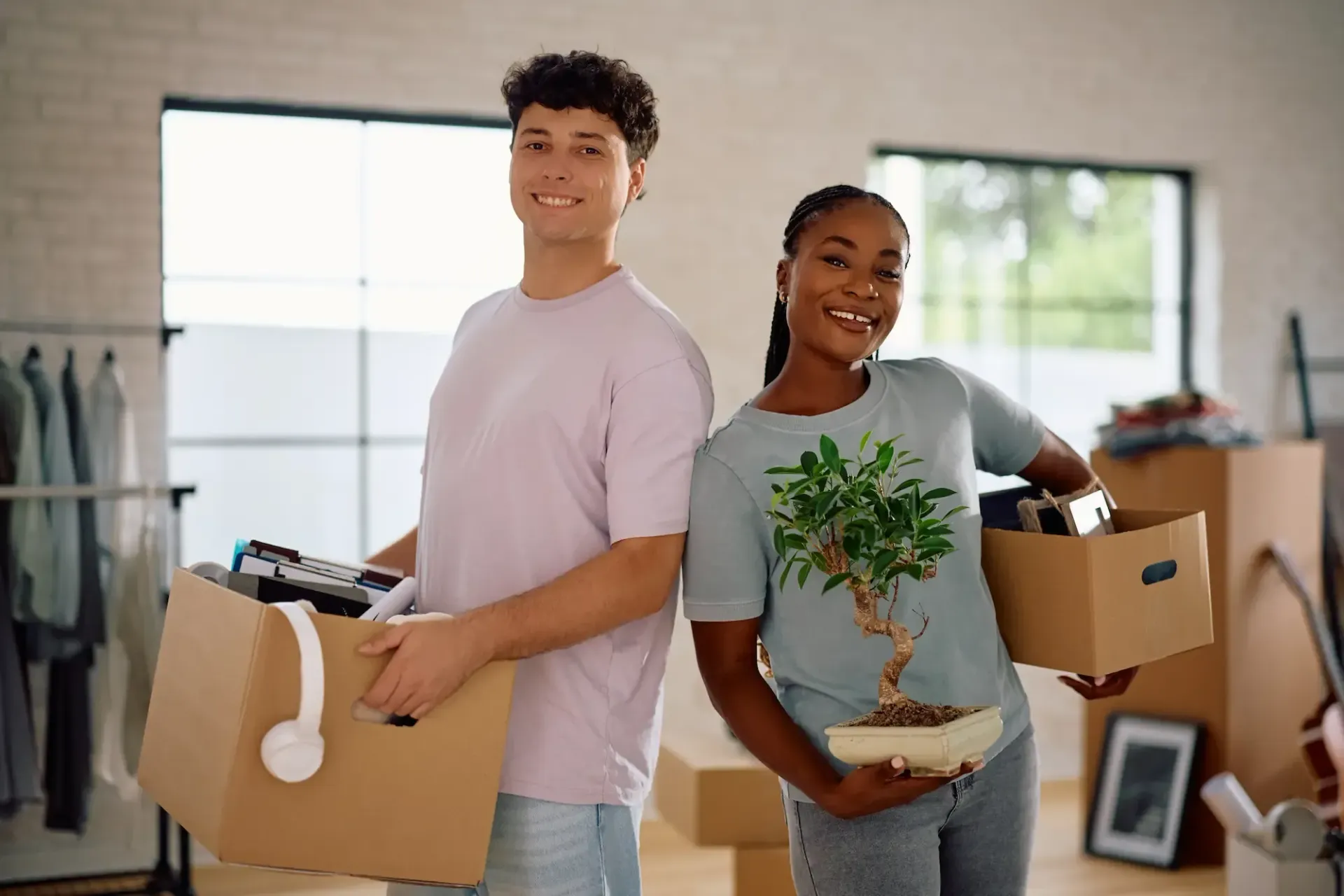 Couple smiling, holding moving boxes in a new home. Man in pink shirt, woman in blue shirt with plant.