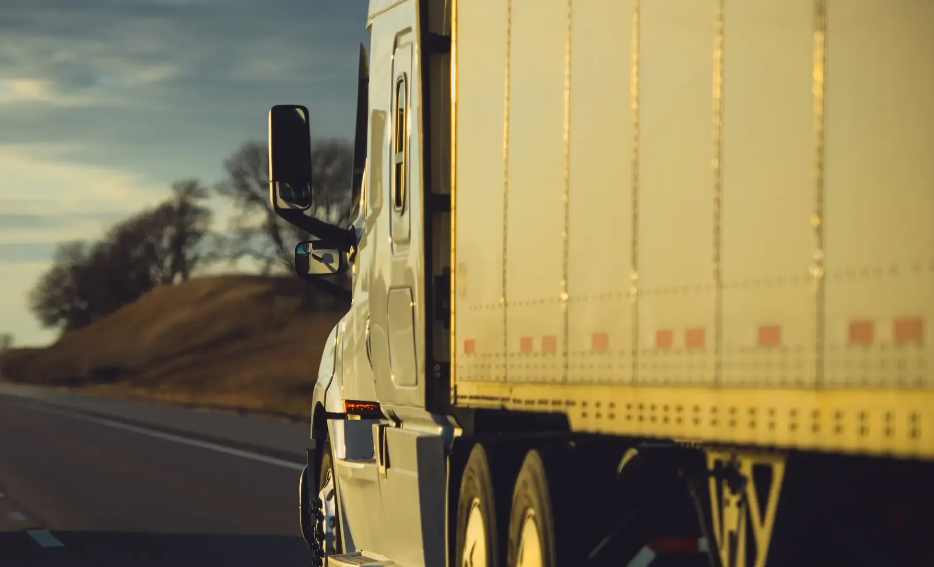 Semi-truck on a highway, side view; golden sunlight, a slight hill in the background.