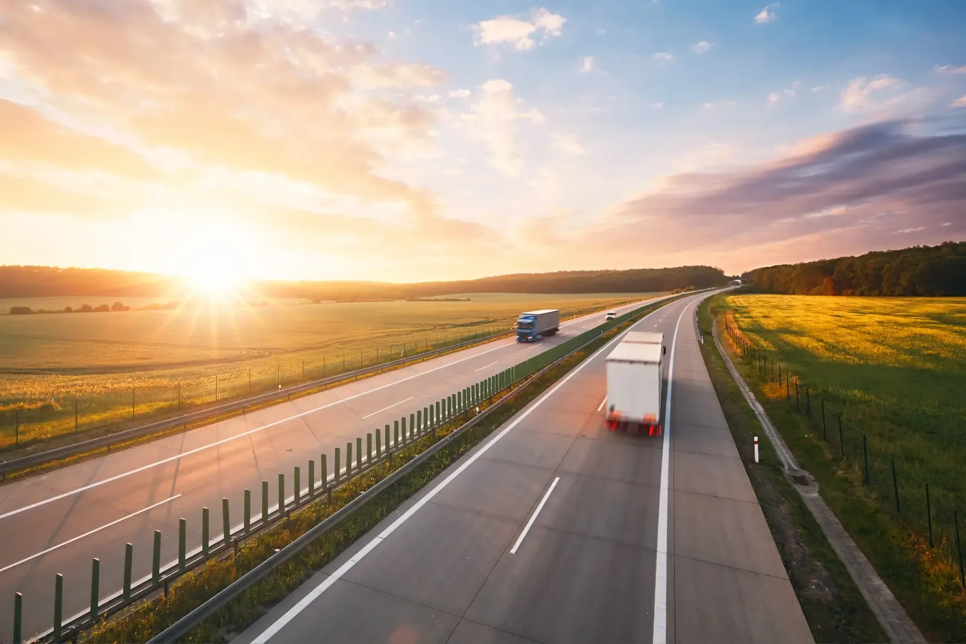 Highway with two semi-trucks driving toward a bright sunset over fields of green.
