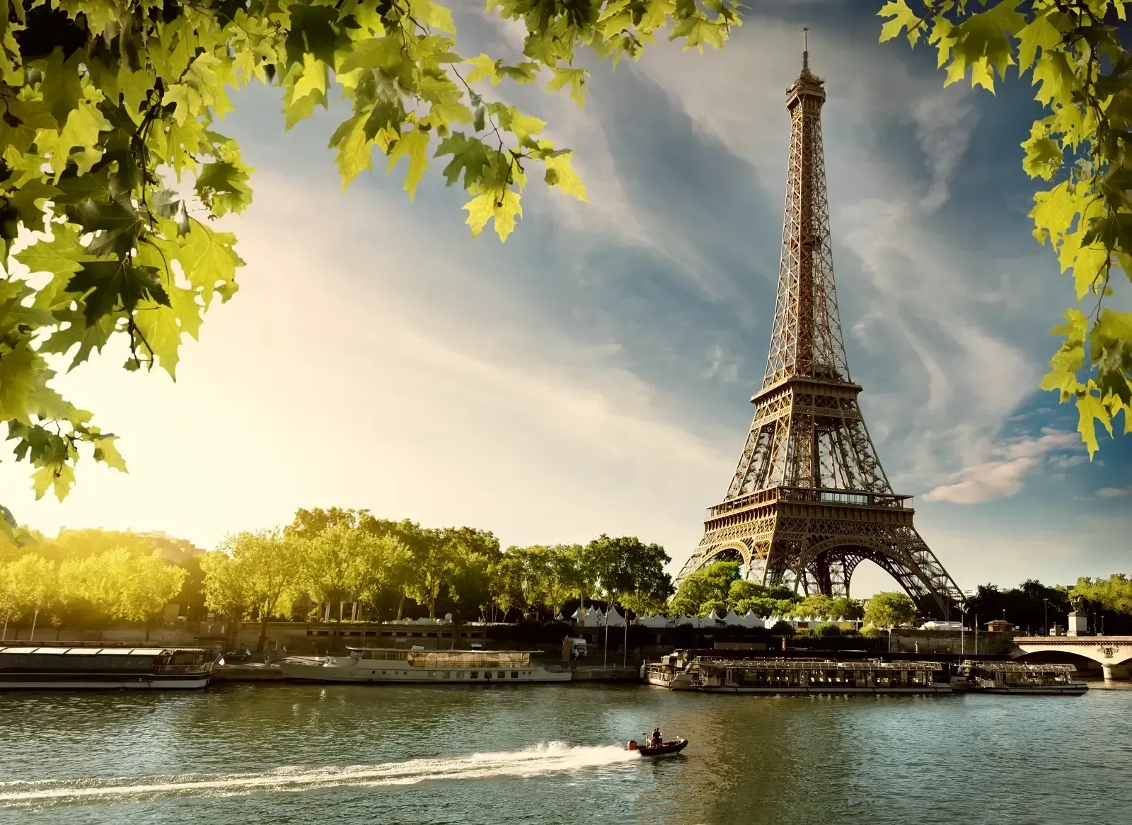The Eiffel Tower rises above the Seine River in Paris, framed by sunlit green leaves with a small boat crossing the water.