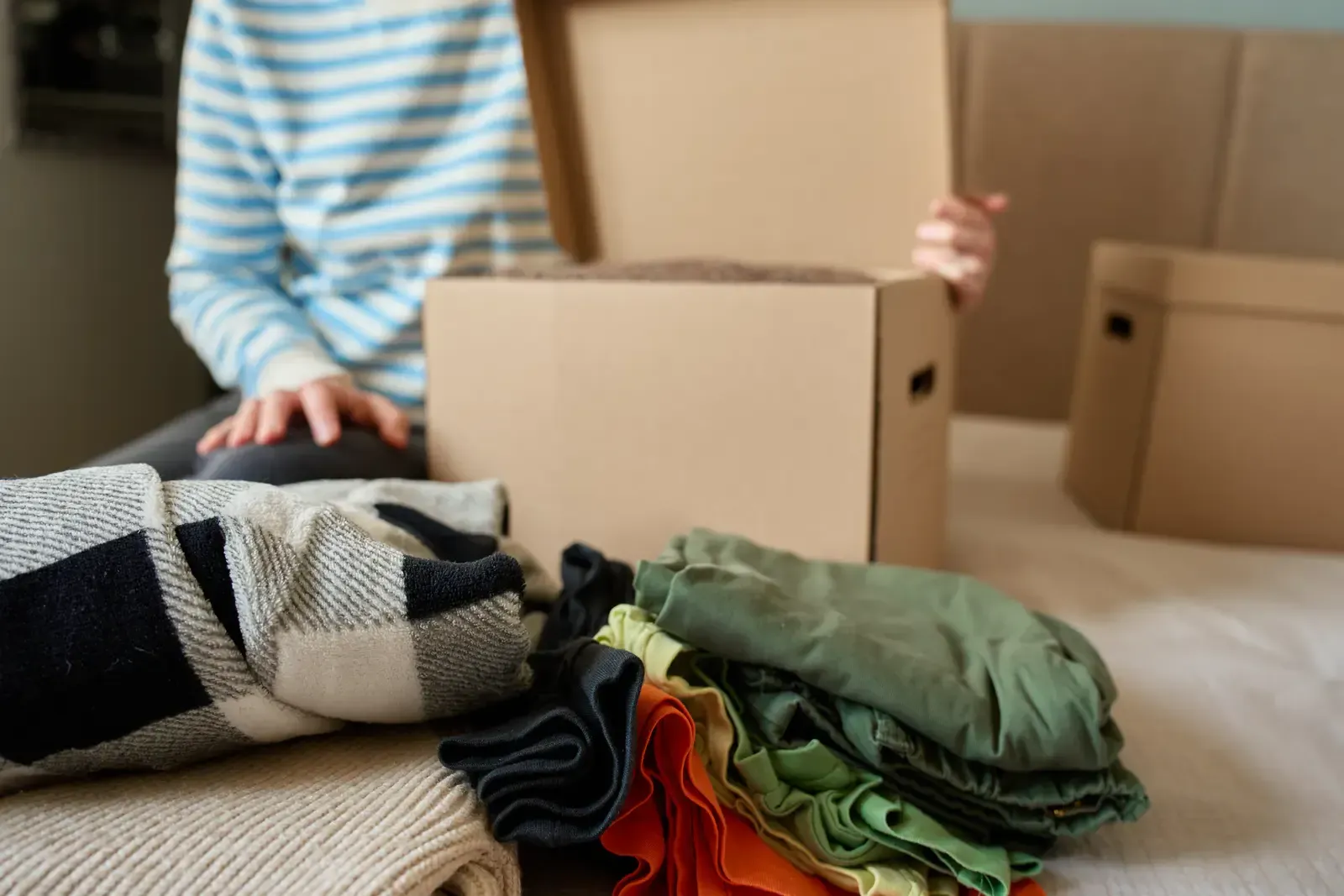 Person packing clothes into a cardboard box on a bed.