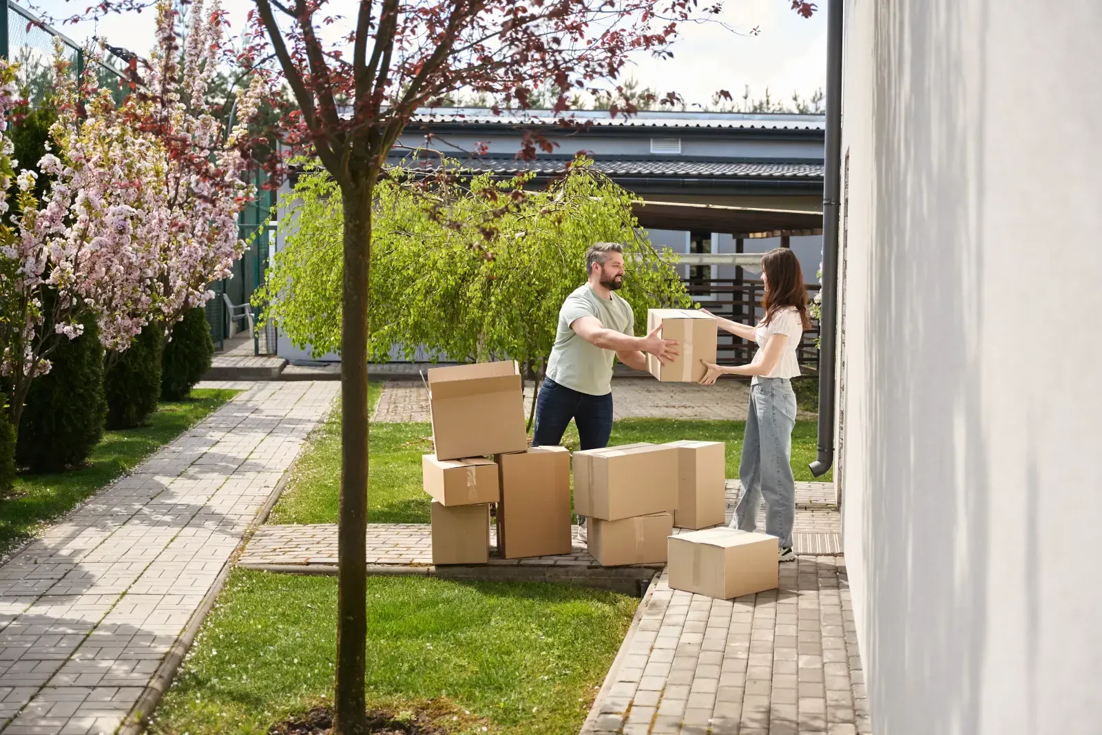 Two people loading cardboard boxes beside a white wall in a sunny yard