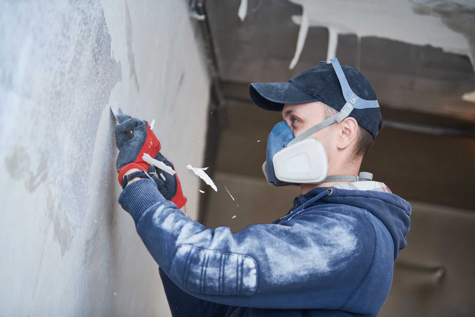 Person wearing a respirator and cap scrapes wall with a tool, possibly removing old material in a dusty indoor setting.