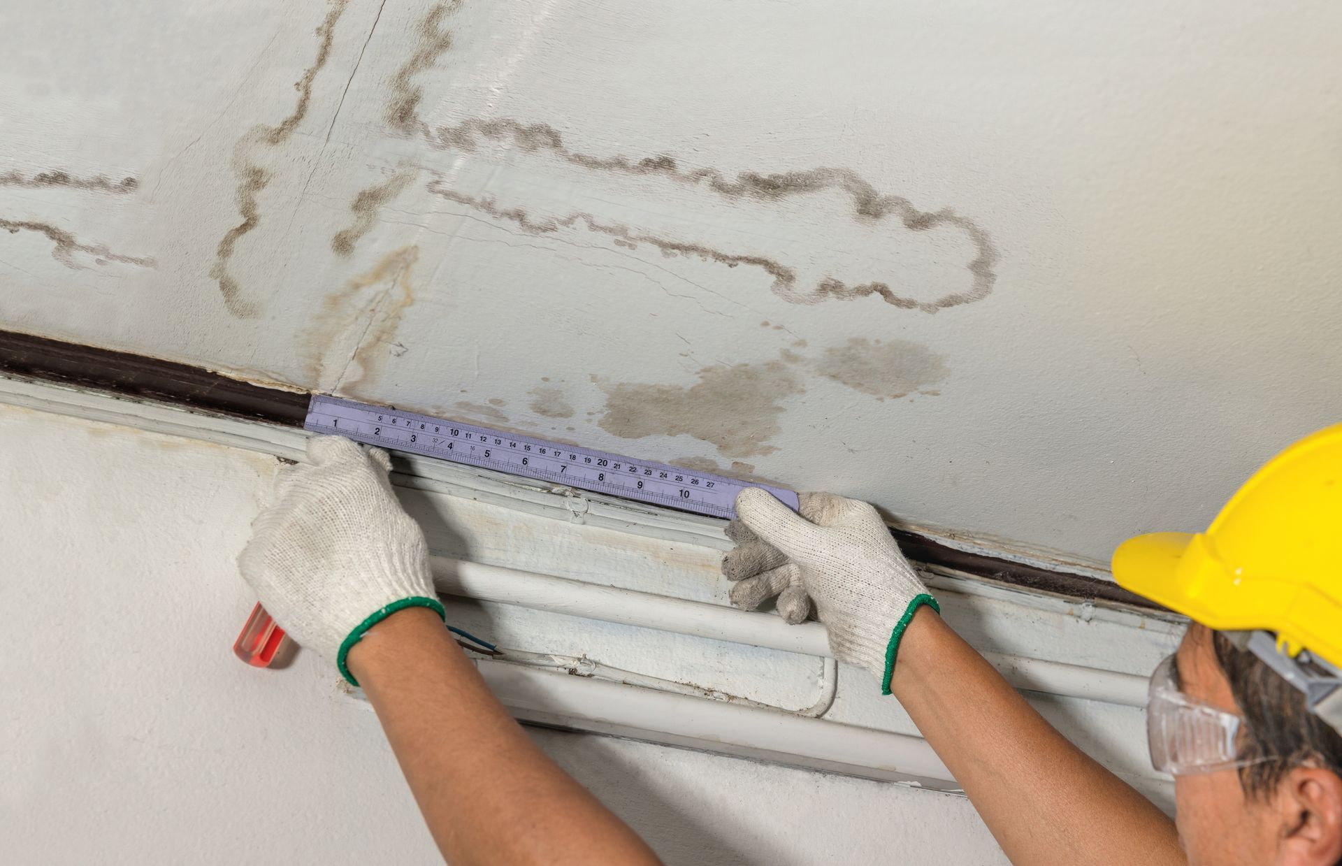 Person in hard hat and gloves applying sealant to water-damaged ceiling. Brown mold stains visible.