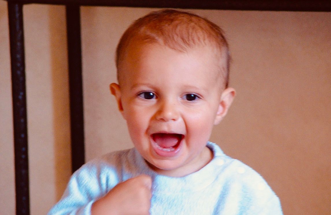 Happy toddler with open mouth and raised arm, wearing a light blue sweater.