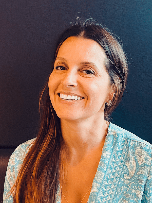 Femme souriante, regardant l'objectif. Elle a de longs cheveux bruns et porte un haut bleu à motifs.