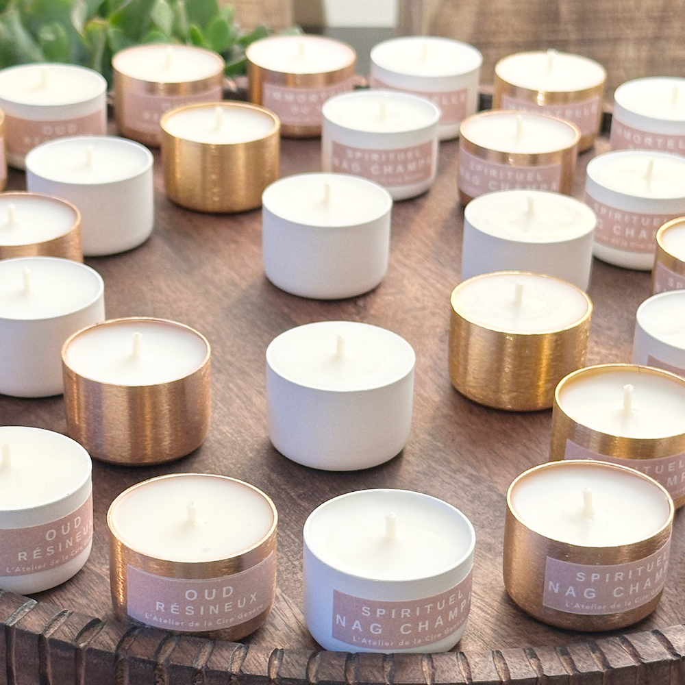 Bougies blanches et dorées sur un plateau en bois sculpté foncé, certaines avec des étiquettes roses.