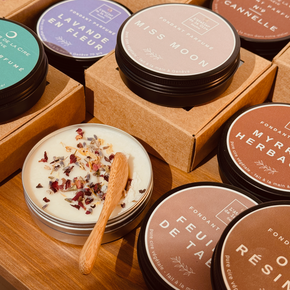 Plusieurs petites boîtes de cire parfumée fondante, avec une spatule en bois et des fleurs séchées, sur une surface en bois.
