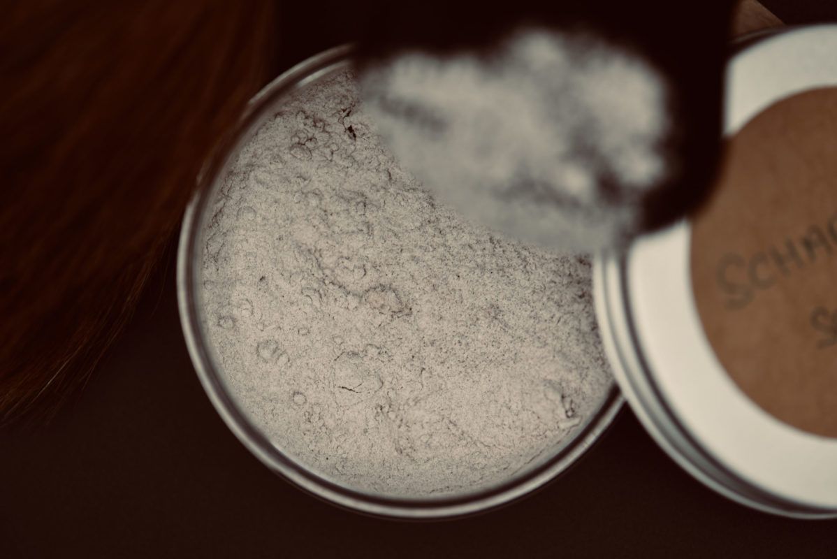 Open tin of light-colored powder with a makeup brush poised above it.
