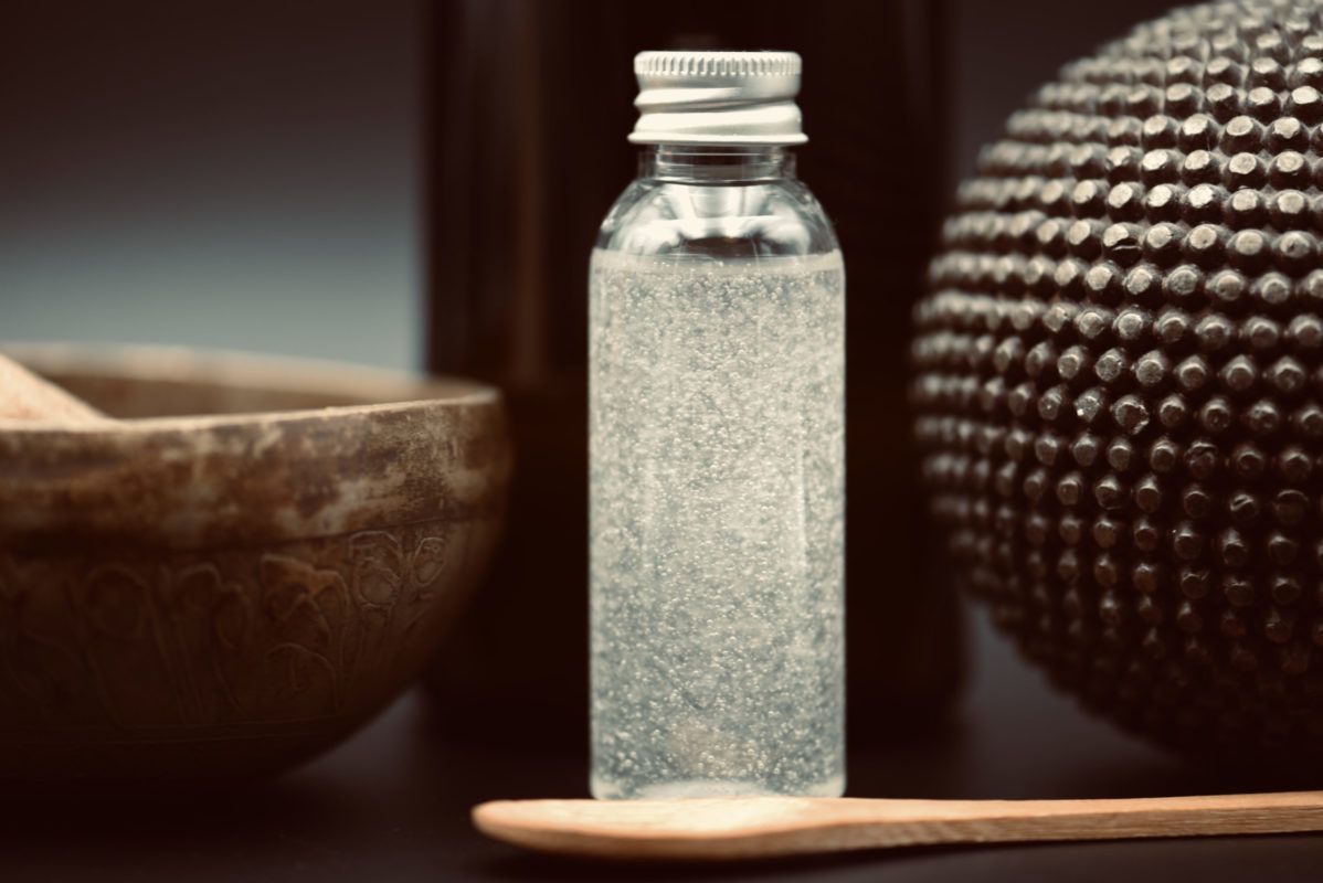 Clear bottle with silver cap filled with liquid, on wooden spoon; bowl and decorative sphere in background.