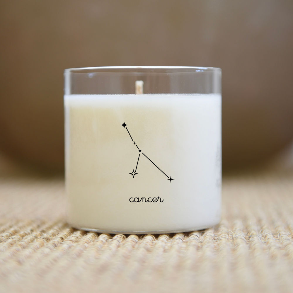 Une bougie dans un verre transparent avec le motif de la constellation du Cancer et le texte « cancer » sur une surface texturée.