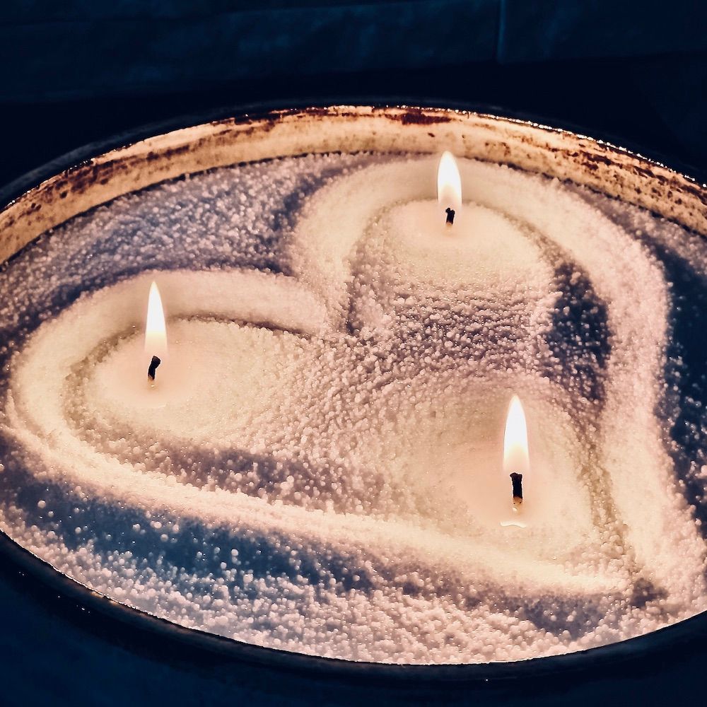 Bougie allumée à trois mèches dans un récipient en verre circulaire, posée sur un fond de cire rose, sur une surface en marbre.